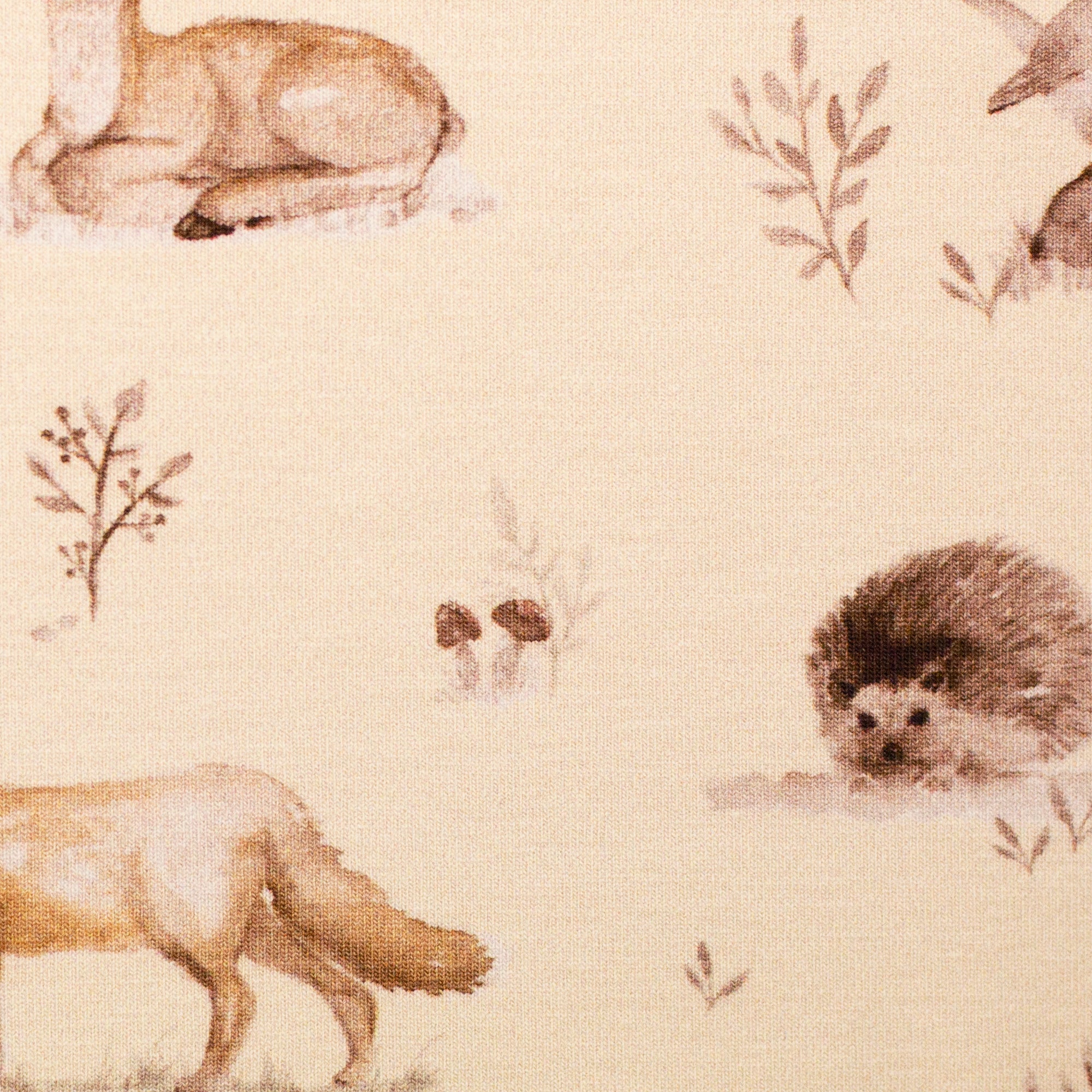 Zu sehen ist eine Nahaufnahme eines weichen, elastischen Baumwolljersey-Stoffs in beige mit Motiven von Waldtieren wie Reh, Fuchs und Hase, der durch seine angenehme, glatte Oberfläche ideal für Kleidung, Accessoires und kreative DIY-Projekte geeignet ist.