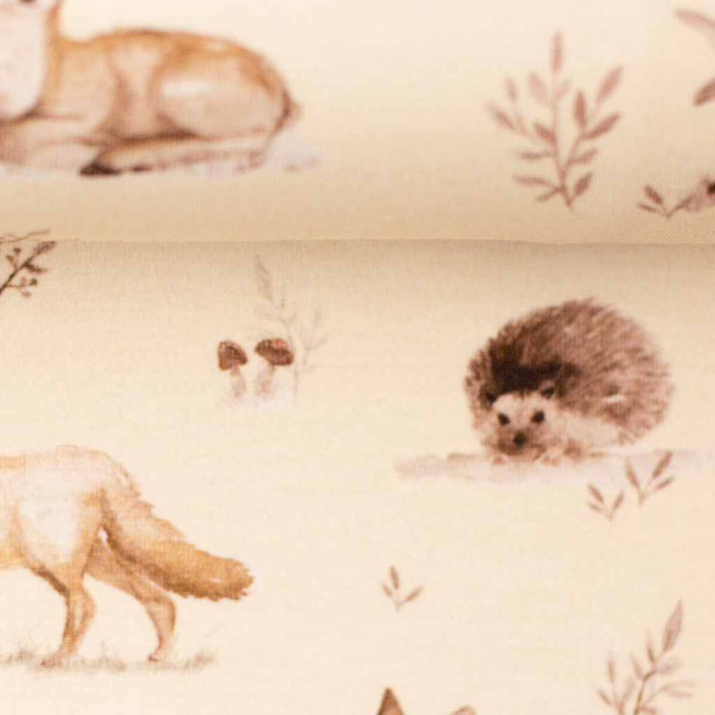 Zu sehen ist eine Nahaufnahme eines weichen, elastischen Baumwolljersey-Stoffs in beige mit Motiven von Waldtieren wie Reh, Fuchs und Hase, der durch seine angenehme, glatte Oberfläche ideal für Kleidung, Accessoires und kreative DIY-Projekte geeignet ist.
