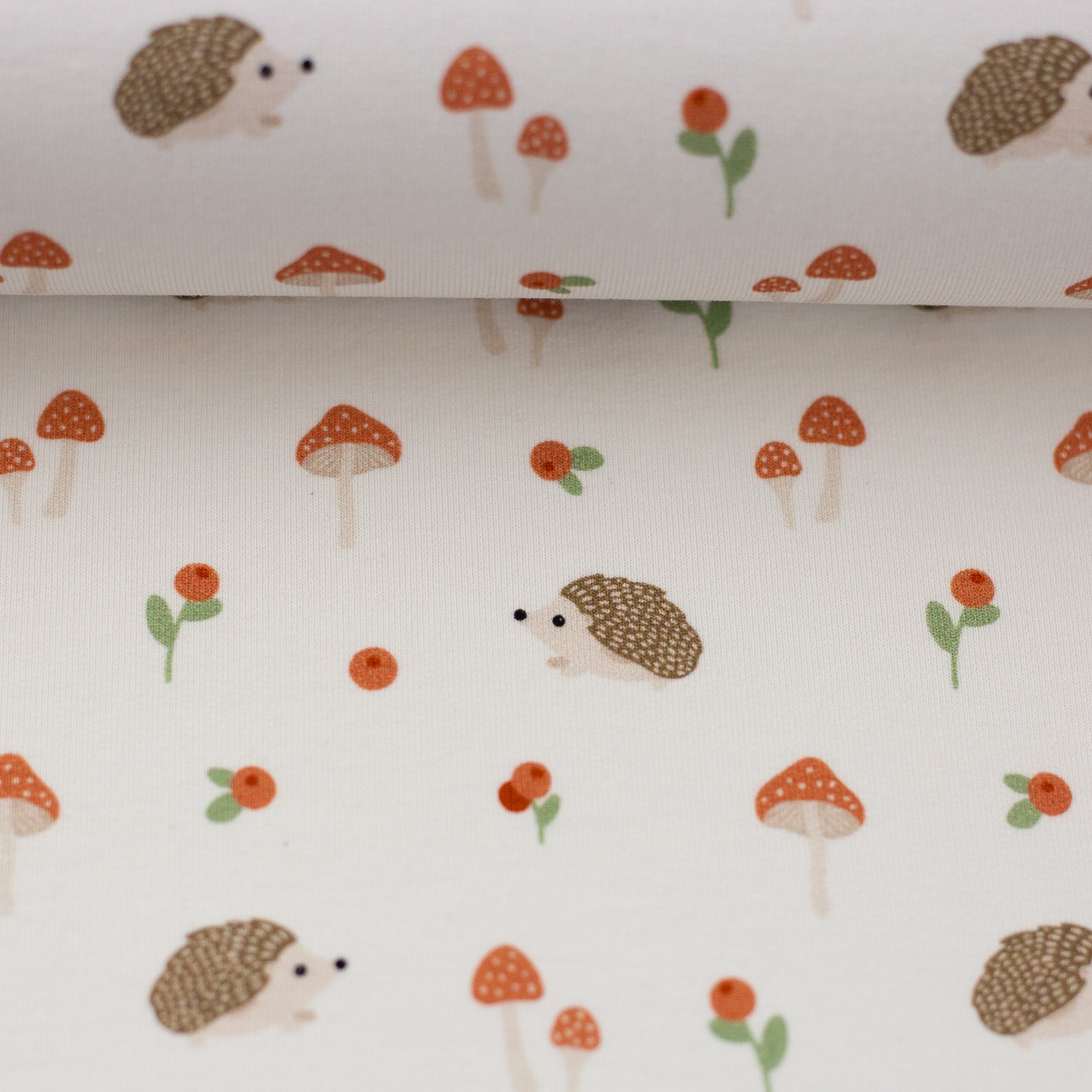 Zu sehen ist eine Nahaufnahme eines weichen, elastischen Baumwolljersey-Stoffs in weiß mit niedlichen Igeln in Naturfarben, der durch seine angenehme, glatte Oberfläche ideal für Kleidung, Accessoires und kreative DIY-Projekte geeignet ist.