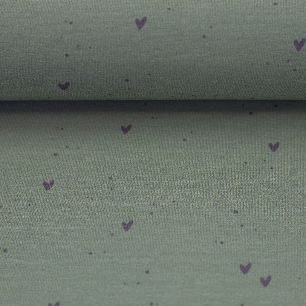 Zu sehen ist eine Nahaufnahme eines weichen, elastischen Baumwolljersey-Stoffs in Grün mit süßen Herz-Motiven, der durch seine angenehme, glatte Oberfläche ideal für Kleidung, Accessoires und kreative DIY-Projekte geeignet ist.
