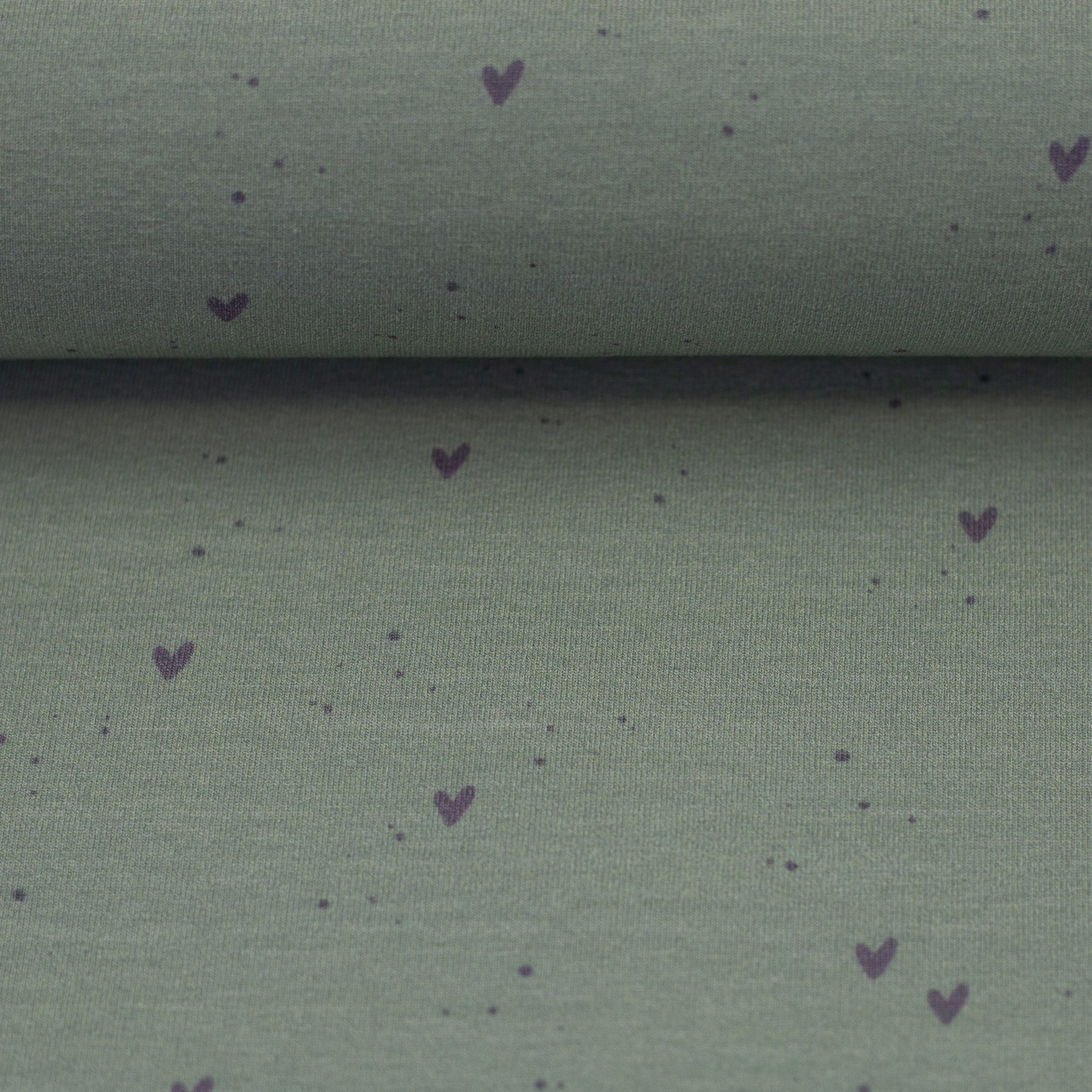 Zu sehen ist eine Nahaufnahme eines weichen, elastischen Baumwolljersey-Stoffs in Grün mit süßen Herz-Motiven, der durch seine angenehme, glatte Oberfläche ideal für Kleidung, Accessoires und kreative DIY-Projekte geeignet ist.